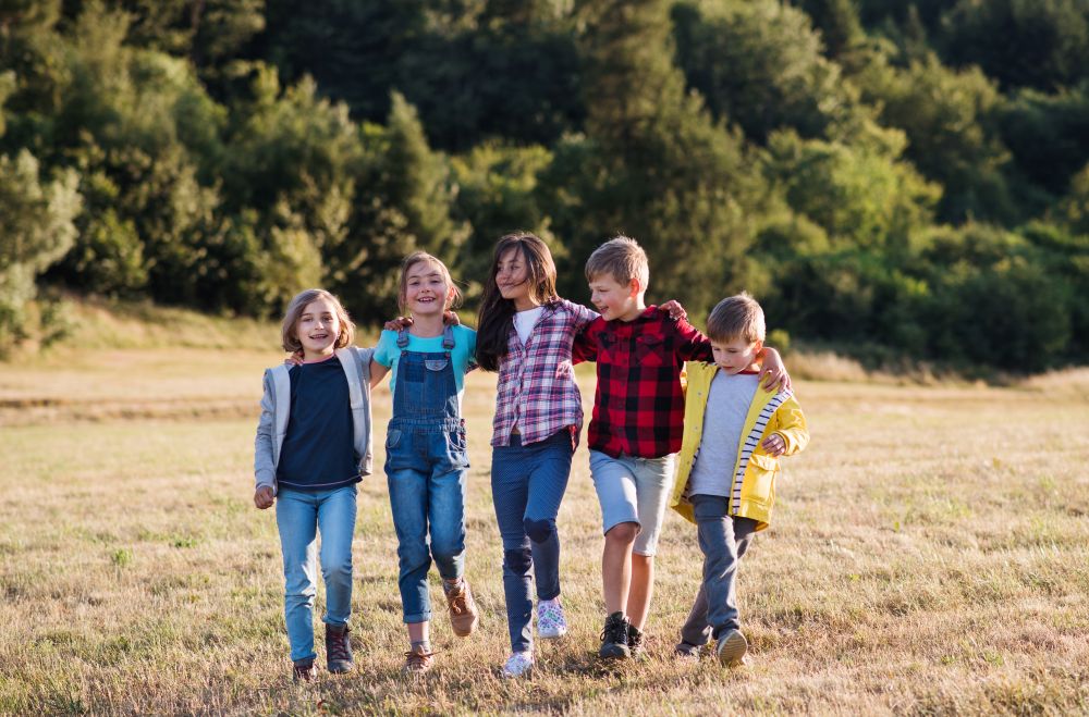 Fünf Kinder gehen über eine gemähte Wiese vor einem Wald, lachen und haben die Hände über die Schultern gelegt.