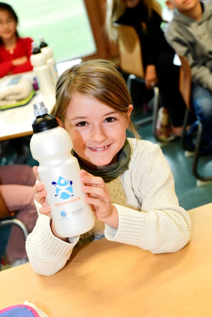 Ein Mädchen sitz an ihrem Schultisch und hält grinsend ihre Wasserschulenflasche in den Händen.