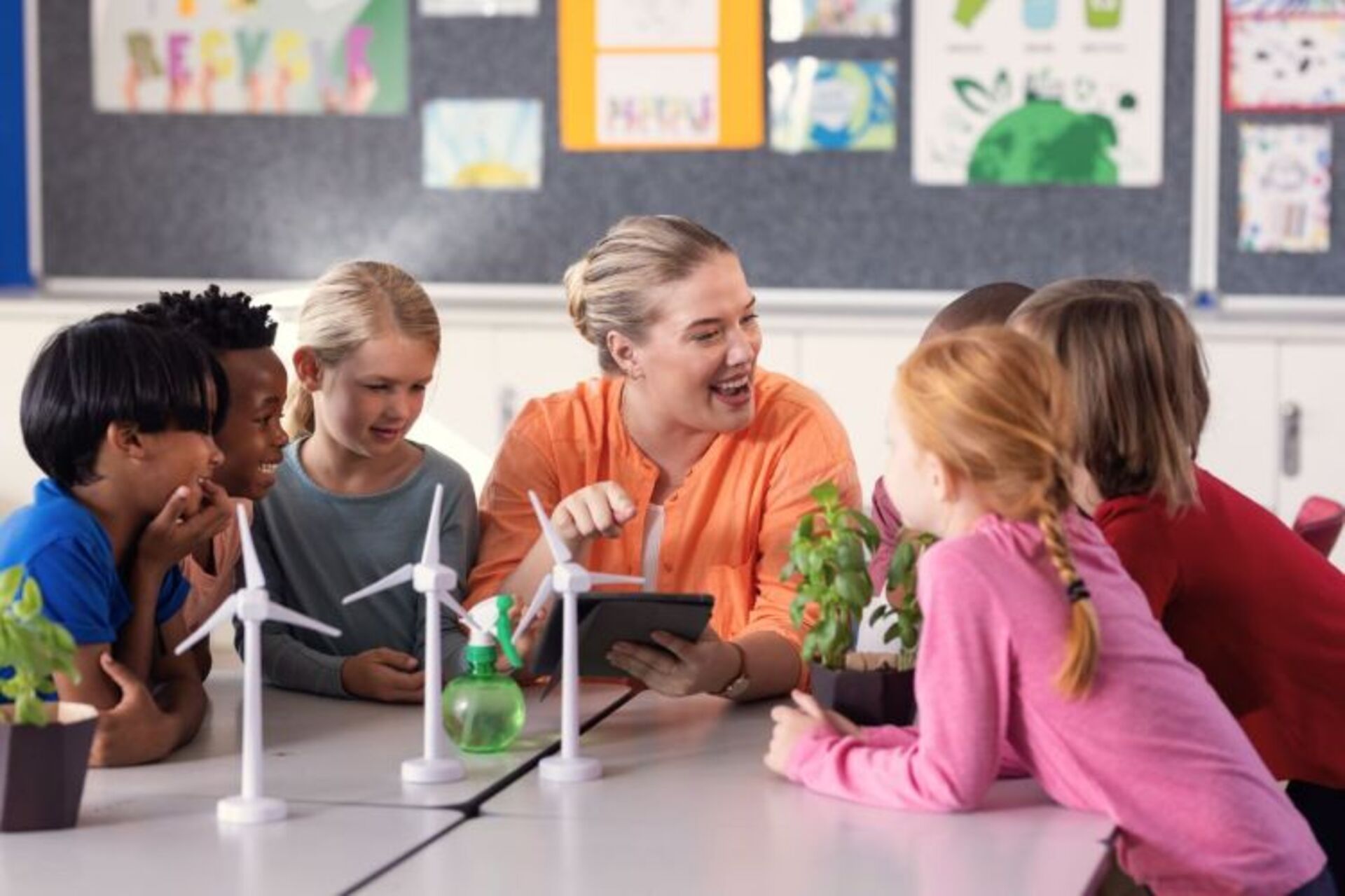 Eine Lehrerin sitzt an einem Tisch mit Windrädern und einem Tablet in der Hand und erklärt den Kindern etwas.