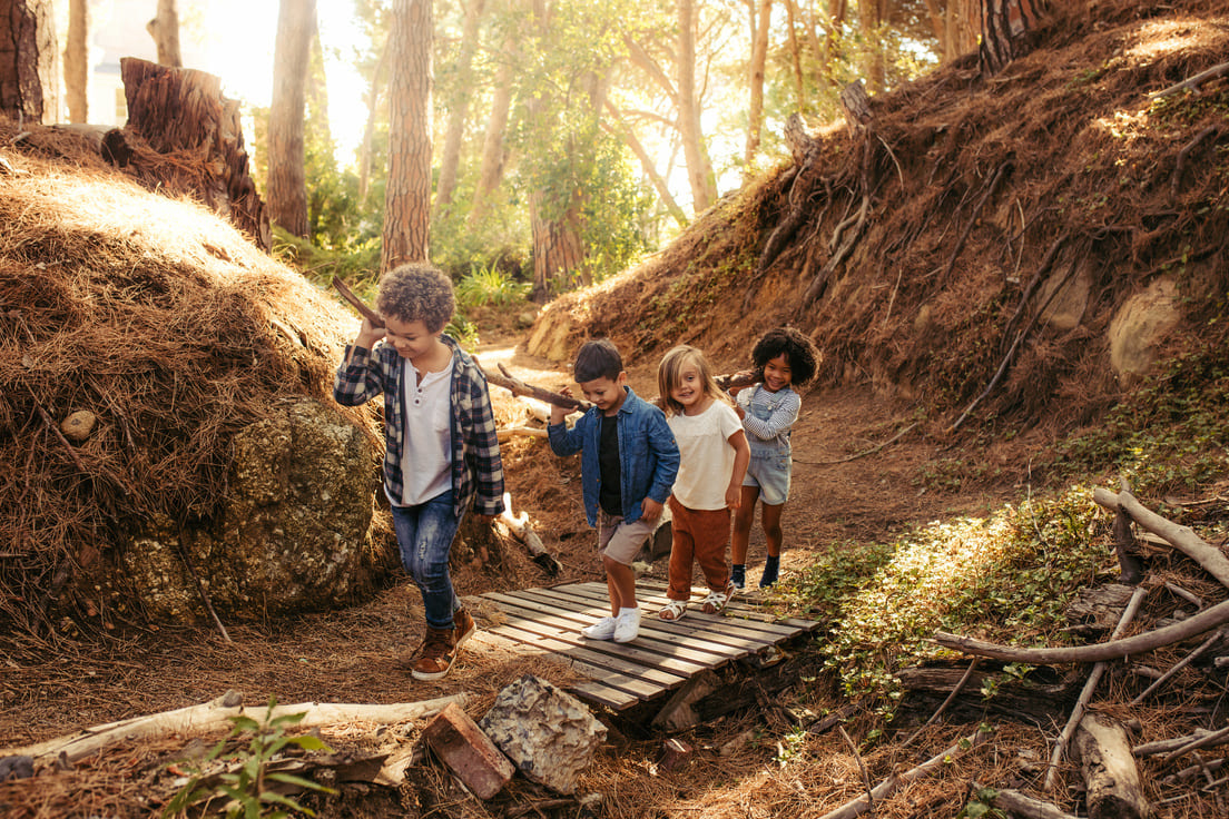 Vier Kinder spazieren über einen Holzsteg im Wald und tragen dabei gemeinsam einen Ast über den Schultern.