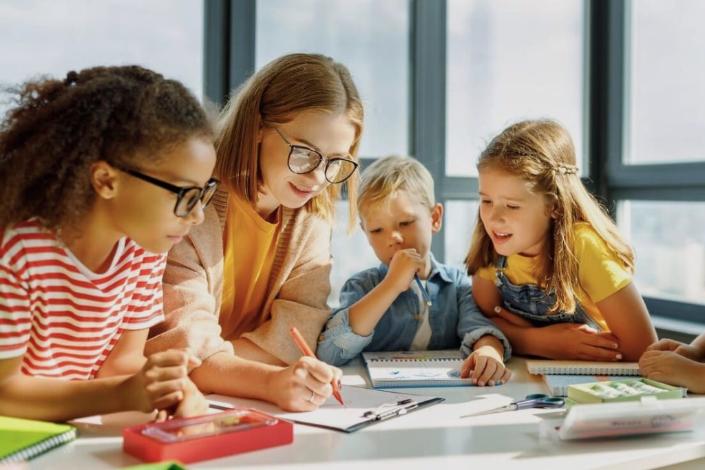 Eine Lehrerin zeichnet etwas auf einem Klemmbrett und drei Kinder schauen ihr dabei zu.