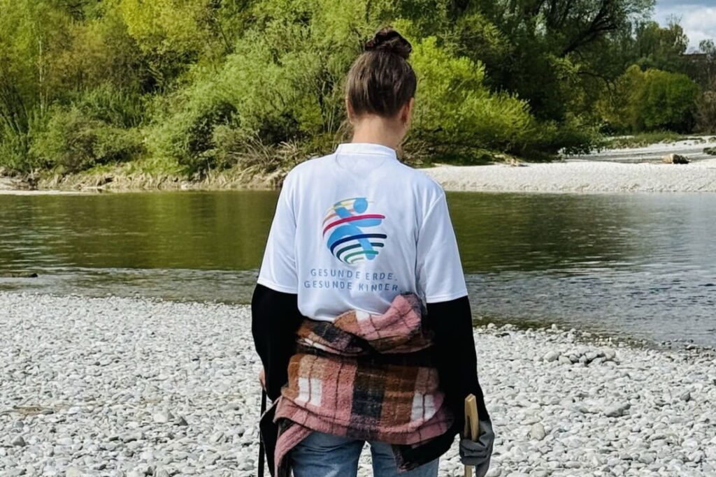 Eine Frau mit einem T-Shirt von Gesunde Erde. Gesunde Kinder. sammelt Müll am Ufer der Isar.