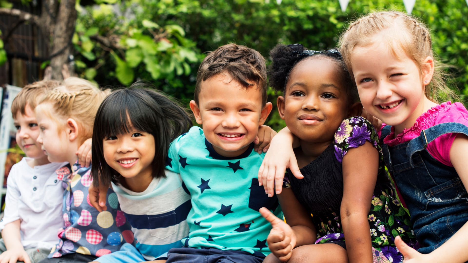 Sechs Kinder sitzen vor grünen Bäumen nebeneinander, haben die Arme umeinander gelegt und lachen.