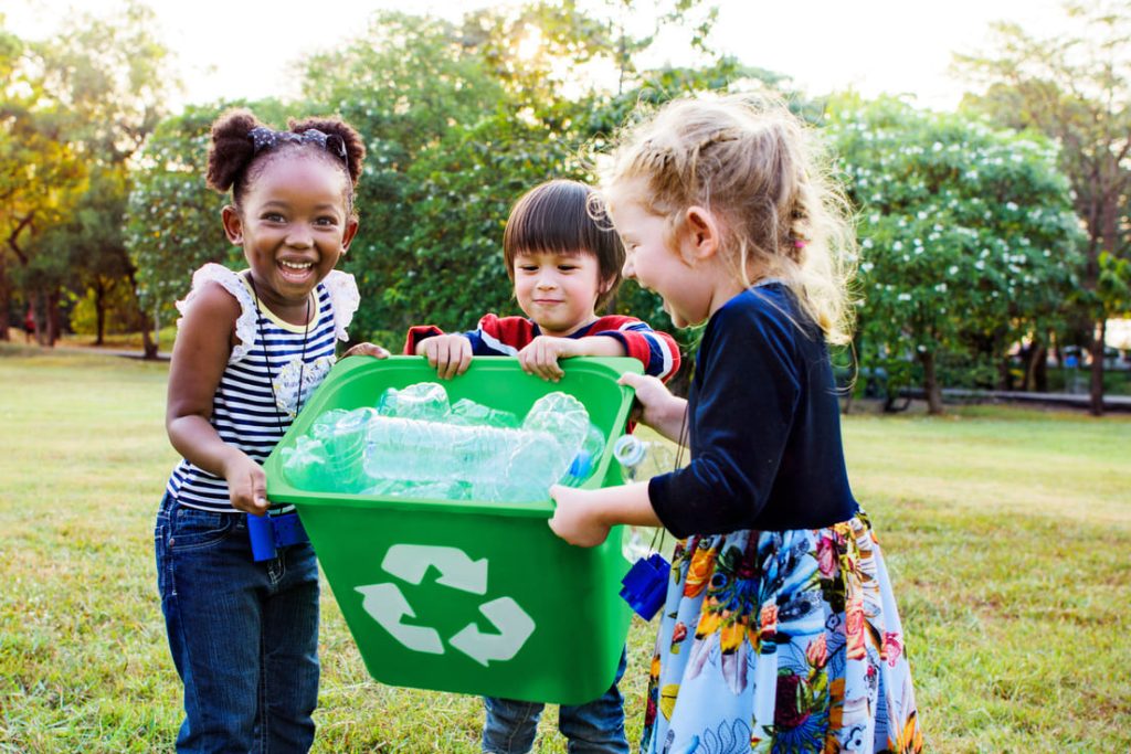 Kinder lachen auf einer Wiese und tragen eine Recyclingkiste mit Plastikflaschen